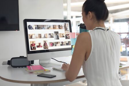 Rear view of Asian female graphic designer drinking water while working on computer at desk in a modern officeの写真素材