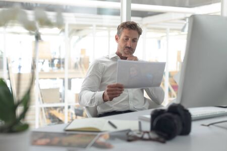Front view of thoughtful Caucasian male graphic designer looking at photograph on desk in a modern officeの写真素材