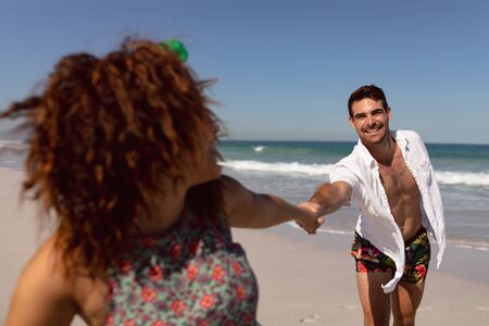 Front view of happy young Mixed-race couple holding hands on beach in the sunshineの写真素材