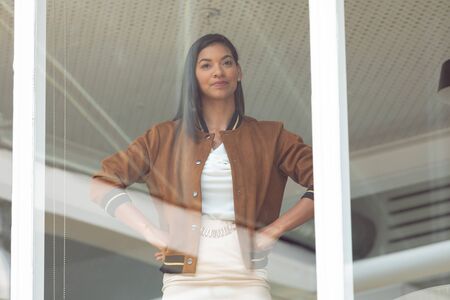 Front view of happy mixed race businesswoman with hands on hips standing near window in a modern officeの写真素材