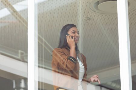Side view of happy mixed race businesswoman talking on mobile phone near window in a modern officeの写真素材