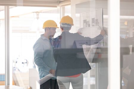 Rear view of Caucasian architects discussing over blueprint on glass board in a modern officeの写真素材