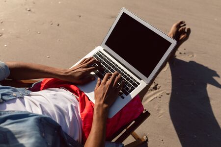 Low section of African-american man using laptop while relaxing in a beach chair on the beacの写真素材