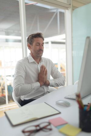 Front view of Caucasian business male executive doing yoga at desk in a modern officeの写真素材