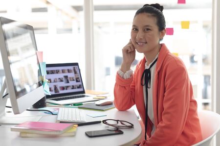 Front view of beautiful Asian female graphic designer looking at camera on desk in a modern office