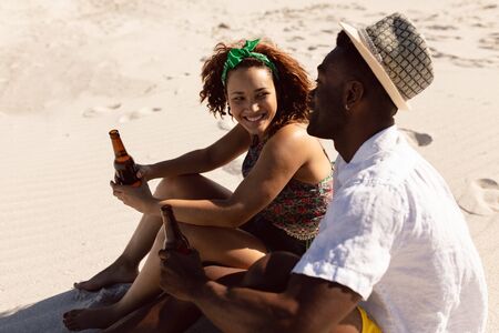 Side view of happy young Mixed-race couple with beer bottle talking with each other on beach in the sunshineの写真素材