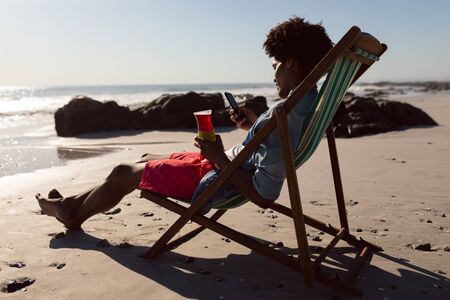 Side view of young African-american man using mobile phone while having cocktail in a beach chairの写真素材