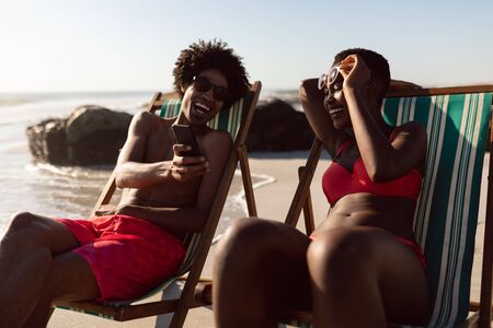 Front view of happy African-american couple having fun on mobile phone while relaxing in a beach chair on the beachの写真素材