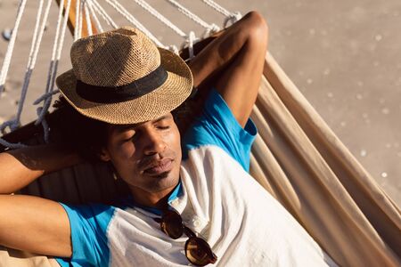 Front view of young African-american man with hat sleeping in a hammock on the beachの写真素材