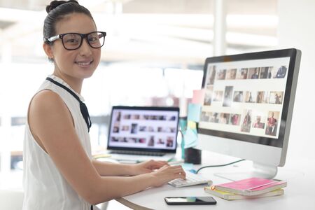 Front view of Asian female graphic designer looking at camera while working on computer at desk in a modern officeの写真素材