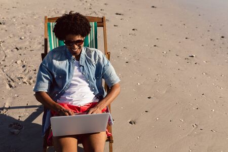 Front view of young African-american man using laptop while relaxing in a beach chair on the beachの写真素材