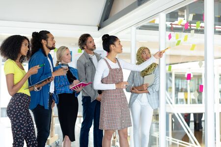 Front view of diverse business people discussing over sticky notes on glass wall in a modern office