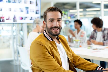 Portrait of Caucasian male fashion designer sitting in the conference room at office. On the background, diverse business people talking around a table in the conference room at office.の写真素材