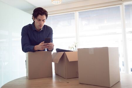 Front view of Caucasian male executive using mobile phone while leaning on cardboard box in office