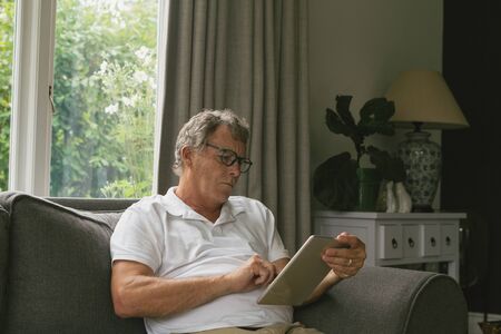 Front view of active senior Caucasian man sitting on sofa and using digital tablet in living room at...
