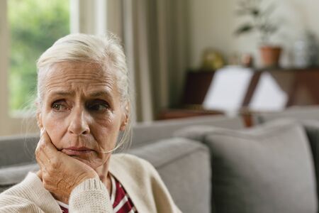 Front view of tensed active senior Caucasian woman with hand on face sitting on sofa in a comfortable homeの写真素材