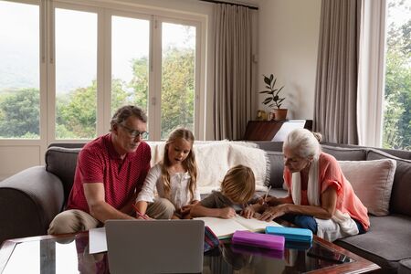 Front view of Caucasian grandparents helping grandchildren with homework in living room at...