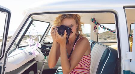 Front view of beautiful Caucasian woman taking photo with digital camera in camper van at beach