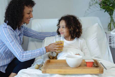 Side view of happy mixed race mother feeding her son orange juice in bed in the ward at hospitalの写真素材