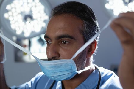 Front view of mature mixed race male surgeon wearing surgical mask in operation room at hospitalの写真素材