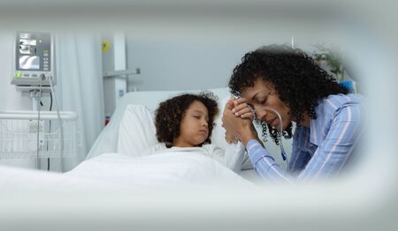 Side view of sad mixed race mother holding hands of her son in the ward at hospital