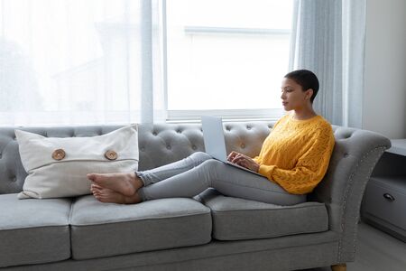 Side view of African american woman using laptop on a sofa in living room at homeの写真素材