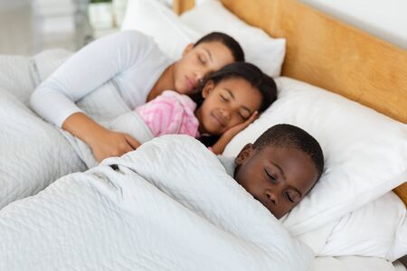 Front view of African american Mother and children sleeping together in bed in bedroom at homeの写真素材