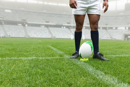 Low section of African American male rugby player standing near rugby ball in stadiumの写真素材