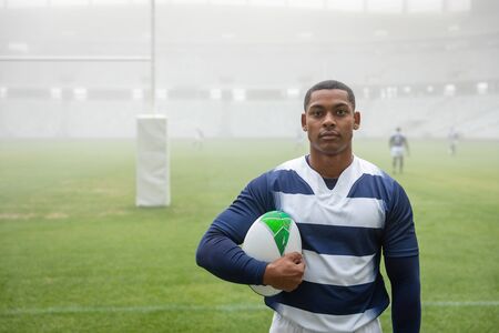 Portrait of African american male rugby player holding a rugby ball in stadium. With players in the...