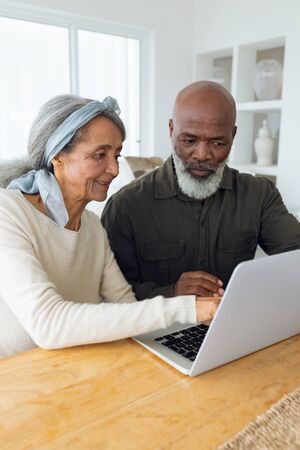 Front view of diverse senior couple using laptop on table inside a room in beach house. Authentic...