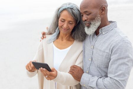Front view of diverse Couple looking at smartphone at the beach. Authentic Senior Retired Life Conceptの写真素材