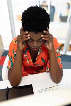 High view of stressed young African american female graphic designer sitting at desk in office. This is a casual creative start-up business office for a diverse teamの写真素材
