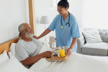 Side view of mixed race healthcare worker talking with old man in bed. Authentic Senior Retired Life...