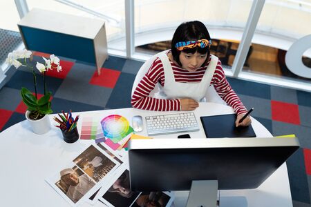 High angle view of beautiful young Asian Female graphic designer using graphic tablet at desk in office. This is a casual creative start-up business office for a diverse teamの写真素材