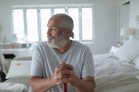 Front view of thoughtful senior Caucasian man sitting on the bed in bedroom. Authentic Senior Retired Life Conceptの写真素材