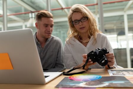 Front view of Caucasian graphic designers discussing over digital camera at desk in office. This is a casual creative start-up business office for a diverse teamの写真素材