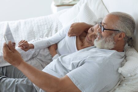 Side view of senior diverse couple lying in bed and watching digital tablet in bedroom. Authentic Senior Retired Life Conceptの写真素材