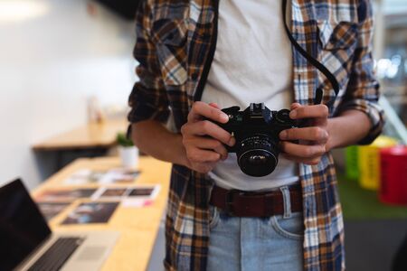 Mid section of mixed-race male graphic designer holding a camera in office. This is a casual creative start-up business office for a diverse teamの写真素材