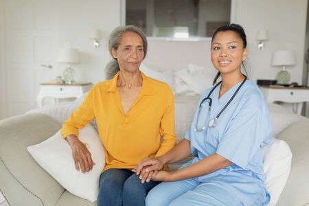 Front view of mixed-race woman healthcare worker and woman sitting on couch indoor. Authentic Senior Retired Life Conceptの写真素材