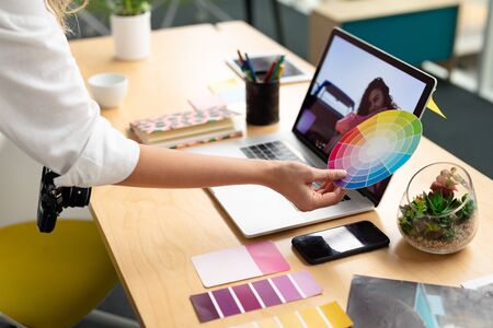 Mid section close-up of Caucasian female graphic designer holding a color swatch at desk in office. This is a casual creative start-up business office for a diverse teamの写真素材