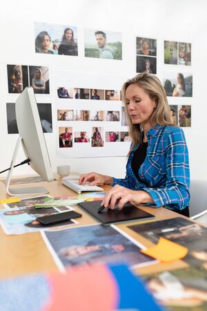 Side view of mature Caucasian female fashion designer using graphic tablet at desk in office. This is a casual creative start-up business office for a diverse teamの写真素材