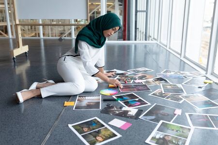 Side view of young mixed race female graphic designer in hijab checking photographs in office. This is a casual creative start-up business office for a diverse teamの写真素材