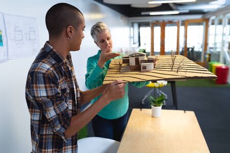 Side view of diverse male and female architects discussing over model structure of house at desk in...