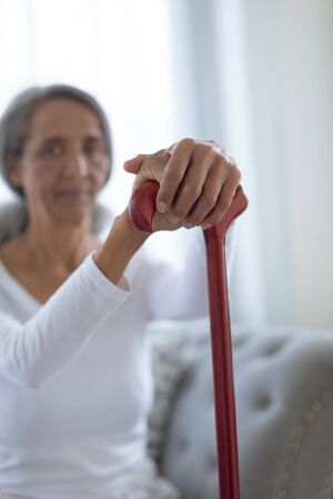 Side view of thoughtful senior mixed race woman sitting on couch while holding a brown walking stick. Authentic Senior Retired Life Conceptの写真素材