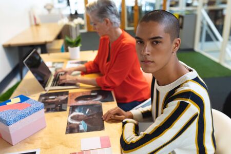 Front view of mixed race male graphic designer looking at camera while Caucasian coworker using...
