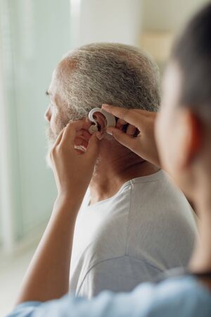 Rear view of healthcare worker putting hearing aid indoor. Authentic Senior Retired Life Conceptの写真素材