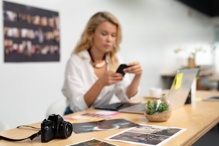 Close-up of digital camera on a desk while female graphic designer using mobile phone in the background at office. This is a casual creative start-up business office for a diverse teamの写真素材