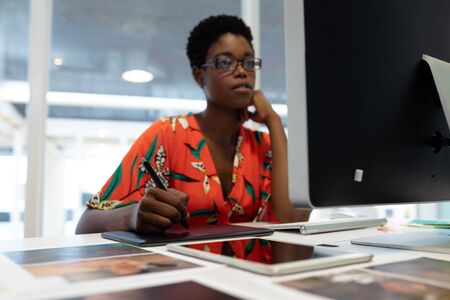 Front view of young African american female graphic designer working on graphic tablet at desk in office. This is a casual creative start-up business office for a diverse teamの写真素材