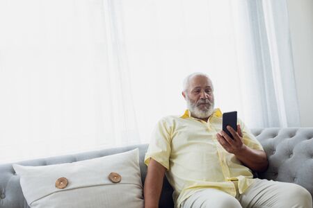 Front view of senior mixed race man using smartphone sitting in a couch. Authentic Senior Retired...