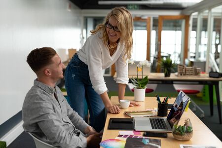 Side view of Caucasian male and female graphic designers interacting with each other at desk in office. This is a casual creative start-up business office for a diverse teamの写真素材